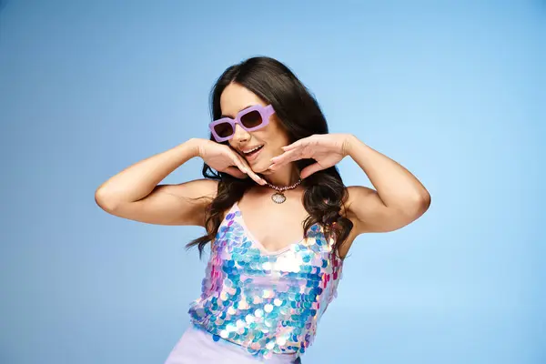 Una mujer con estilo en gafas de sol y una camiseta de lentejuelas logra una pose segura en un estudio de fotografía sobre un vibrante fondo azul. - foto de stock