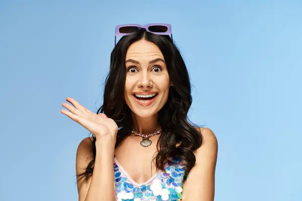 A pretty woman in a bathing suit joyfully raises her hands in a studio, wearing sunglasses against a blue background. — Stock Photo