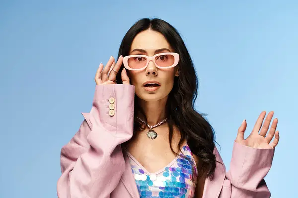 Une femme à la mode dégage des vibrations estivales dans une veste rose et des lunettes de soleil sur fond de studio bleu vif. — Photo de stock