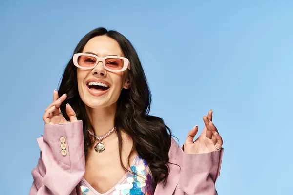 Une femme élégante avec des lunettes de soleil roses et une veste pose en toute confiance dans un studio sur un fond bleu estival. — Photo de stock