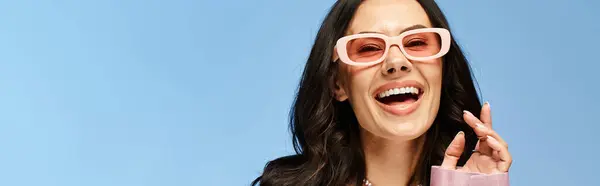 Красива жінка в рожевих сонцезахисних окулярах в студії на синьому фоні, робить комічний вираз. — стокове фото