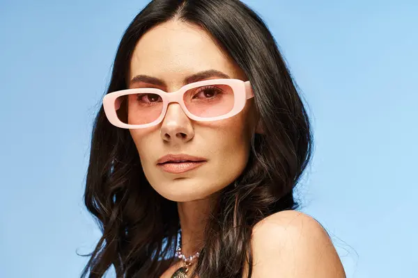 Une femme élégante avec des lunettes de soleil roses pose en toute confiance, respirant les vibrations estivales dans un studio sur un fond bleu. — Photo de stock