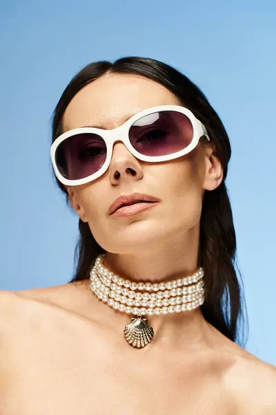 Una mujer elegante con gafas de sol y un collar posa en un estudio sobre un fondo azul brillante, exudando estilo veraniego y sofisticación. - foto de stock