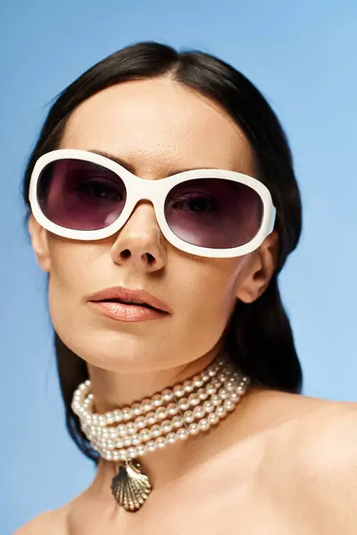 A stylish woman wearing sunglasses and a pearl necklace poses in a studio against a blue background, exuding a glamorous summer vibe. — Stock Photo