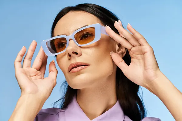 Una mujer de moda con gafas de sol y una camisa púrpura posando con confianza en un estudio con un fondo azul, exudando vibraciones veraniegas. - foto de stock