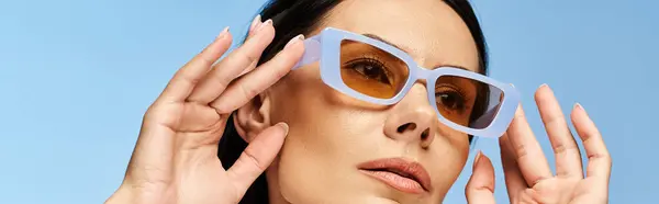 Uma mulher elegante em óculos de sol com as mãos no rosto, exalando frescura em um fundo de estúdio azul. — Fotografia de Stock