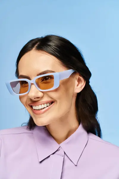 Стильная женщина в солнечных очках и в фиолетовой рубашке позирует в студии на синем фоне, источая летнее очарование. — стоковое фото
