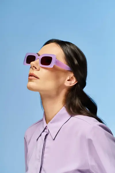 Une femme à la mode en chemise violette et lunettes de soleil pose en toute confiance dans un studio sur fond bleu. — Photo de stock