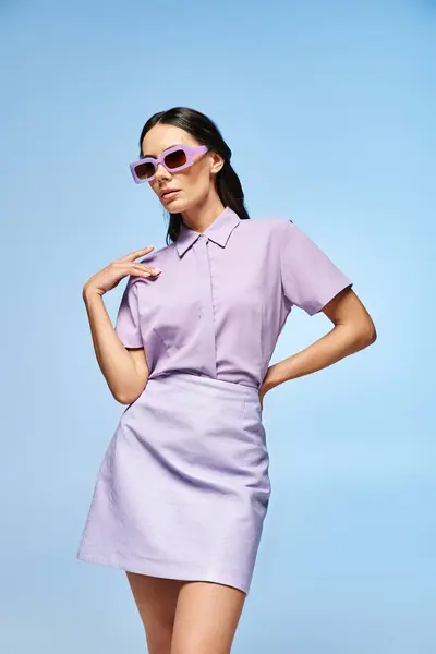 Une femme à la mode se tient en confiance dans une robe courte pourpre et des lunettes de soleil à la mode sur un fond bleu vif. — Photo de stock