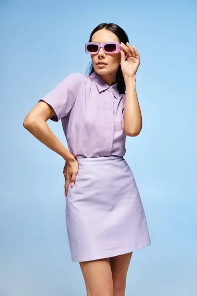 Mujer con estilo en vestido púrpura y gafas de sol posa con confianza en un estudio sobre un fondo azul vibrante. - foto de stock