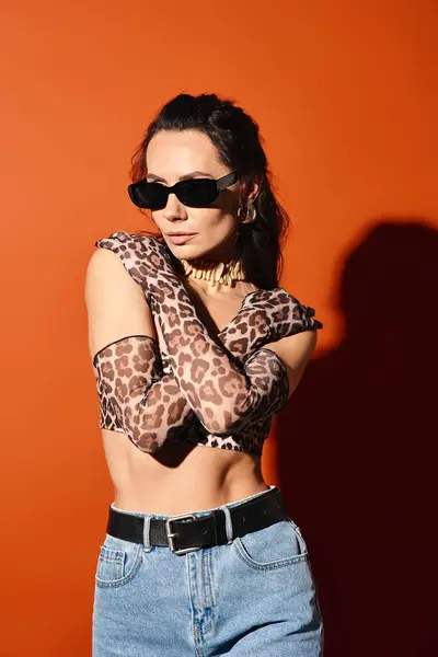 Uma mulher elegante posa no topo de impressão de leopardo e óculos de sol em um fundo laranja, exalando chique de verão. — Fotografia de Stock