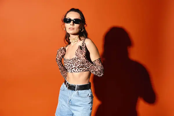 Una mujer con estilo en gafas de sol posa en una parte superior de impresión de leopardo y pantalones cortos de mezclilla sobre un fondo naranja, exudando vibraciones de moda de verano. - foto de stock