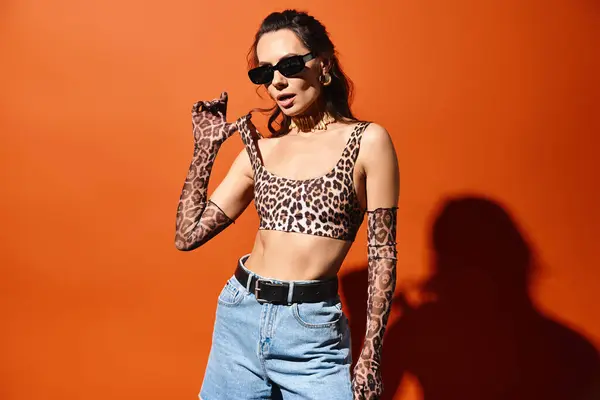 Mulher elegante em óculos de sol posando confiantemente em um topo de impressão de leopardo e jeans contra um fundo laranja. — Fotografia de Stock