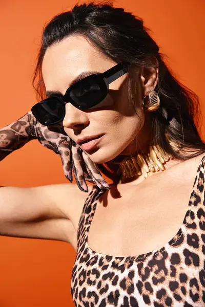 Uma mulher elegante em uma parte superior de impressão de leopardo e óculos de sol posa confiantemente em um estúdio contra um fundo laranja. — Fotografia de Stock