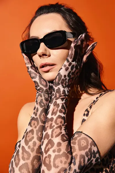 Une femme élégante dégage des vibrations estivales alors qu'elle pose dans des gants imprimés léopard et des lunettes de soleil à la mode sur un fond orange. — Photo de stock