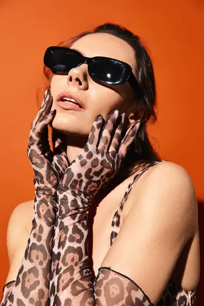 Una mujer elegante adornada con gafas de sol y una bufanda estampada de leopardo irradia moda veraniega en un estudio sobre un fondo naranja. - foto de stock