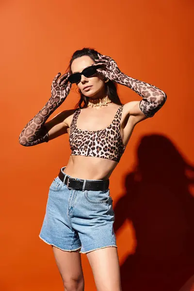 Una donna alla moda in un top stampa leopardo e pantaloncini di jeans trasuda fiducia in un ambiente studio su uno sfondo arancione. — Foto stock