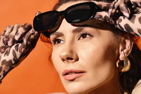 Una donna elegante emana vibrazioni estive mentre indossa con fiducia occhiali da sole e una sciarpa chic sulla testa su uno sfondo arancione. — Foto stock