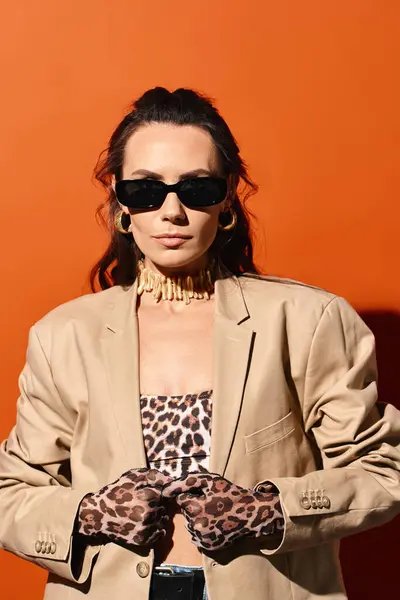 Una mujer de moda con gafas de sol posa en una chaqueta bronceada y guantes estampados de leopardo sobre un fondo de estudio naranja. - foto de stock