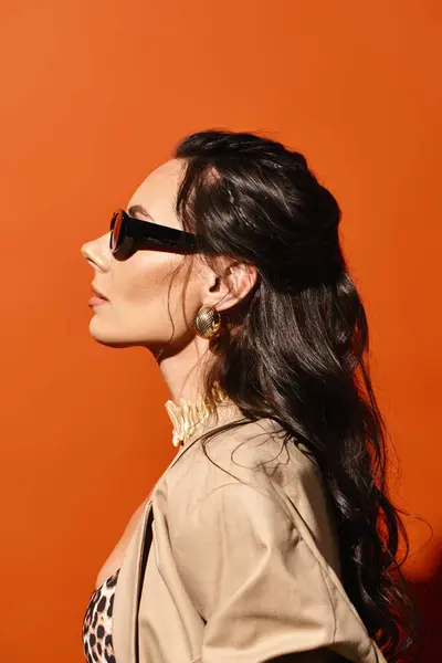 A stylish woman with sunglasses poses confidently in a fashionable jacket against an orange backdrop. — Stock Photo