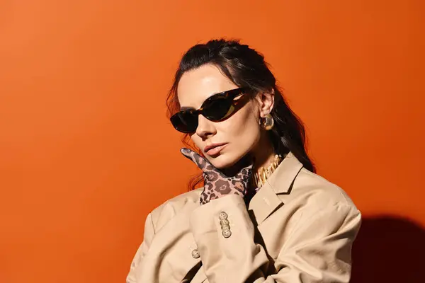 A stylish woman in sunglasses and a trench coat poses confidently against a vibrant orange studio backdrop, exuding summertime chic. — Stock Photo