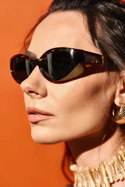 A fashionable woman exudes summer vibes with sunglasses and a gold necklace against an orange backdrop. — Stock Photo