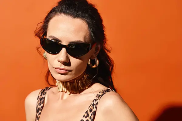 Uma mulher na moda usando óculos de sol e um top de impressão de leopardo posa com confiança em um estúdio contra um fundo laranja. — Fotografia de Stock