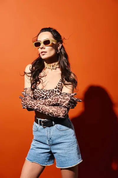 Una mujer elegante con top estampado de leopardo y pantalones cortos de mezclilla, con vibraciones veraniegas exudantes, sobre un fondo naranja. - foto de stock