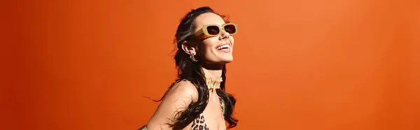 A stylish woman in a leopard print dress and sunglasses poses confidently in a studio against an orange background. — Stock Photo