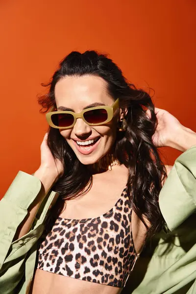 A stylish woman in a leopard print bikini top and sunglasses poses in a summertime fashion shoot against an orange backdrop. — Stock Photo