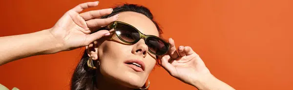 Стильна жінка в сонцезахисних окулярах тримає руки на обличчі, спираючись в теплі, на помаранчевий фон. — стокове фото