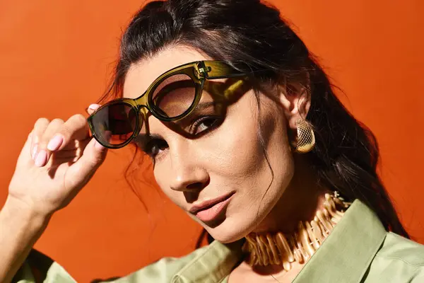 Una donna alla moda che emana vibrazioni estive in una giacca verde elegante e occhiali da sole alla moda sullo sfondo di uno studio arancione. — Foto stock