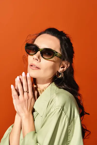 Модна жінка в сонцезахисних окулярах і зеленій сорочці впевнено позує в студії з помаранчевим фоном. — стокове фото