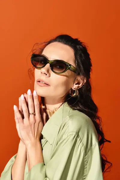 Una mujer de moda con gafas de sol y una camisa verde posa con confianza en un estudio con un fondo naranja. - foto de stock