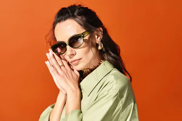 Una donna alla moda che indossa occhiali da sole e una camicia verde posa con fiducia in uno studio su uno sfondo arancione, trasudando chic estivo. — Foto stock
