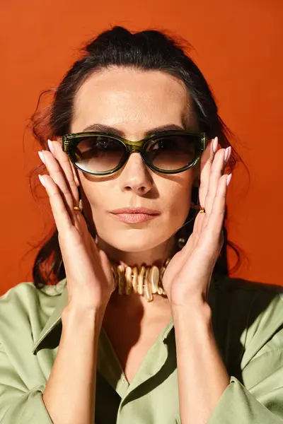 Une femme élégante avec des lunettes de soleil, touchant doucement son visage, respirant confiance et élégance dans un studio lumineux sur fond orange. — Photo de stock
