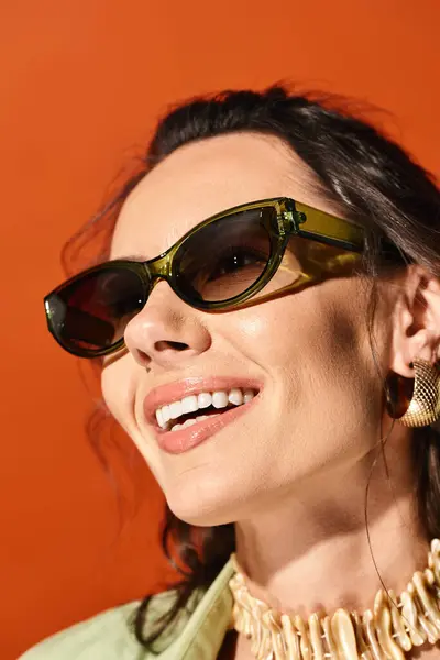 Eine modische Frau strahlt sommerlichen Chic in Sonnenbrille und Statement-Halskette vor orangefarbener Studiokulisse aus. — Stockfoto