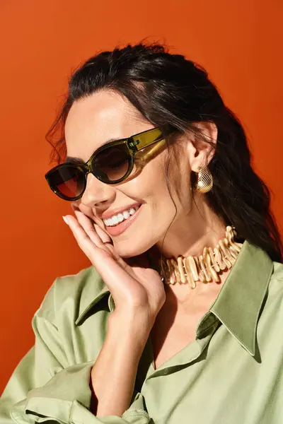 Une femme élégante avec des lunettes de soleil pose en toute confiance dans une chemise verte sur fond de studio orange. — Photo de stock