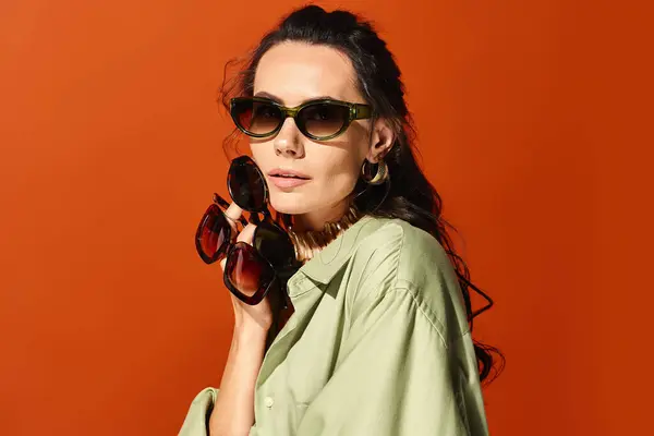 Una donna alla moda in camicia verde e occhiali da sole alla moda posa in uno studio su uno sfondo arancione, trasudando vibrazioni estive. — Foto stock