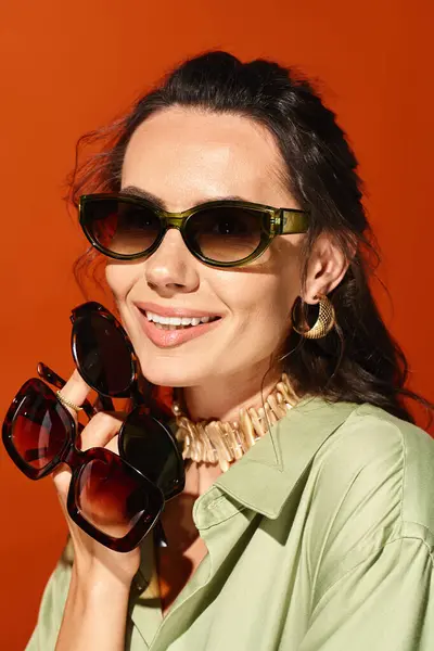 Une femme élégante dans une chemise verte et des lunettes de soleil pose dans un studio sur un fond orange, mettant en valeur la mode estivale. — Photo de stock