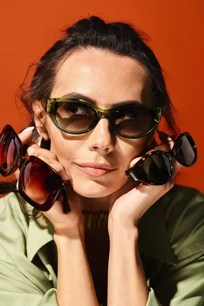 Une femme élégante portant des lunettes de soleil avec ses mains sur son visage, respirant la confiance et les vibrations estivales sur un fond de studio orange. — Photo de stock