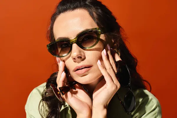 Une femme élégante avec des lunettes de soleil et une chemise verte pose dans un studio avec un fond orange vif, respirant les vibrations de la mode estivale. — Photo de stock
