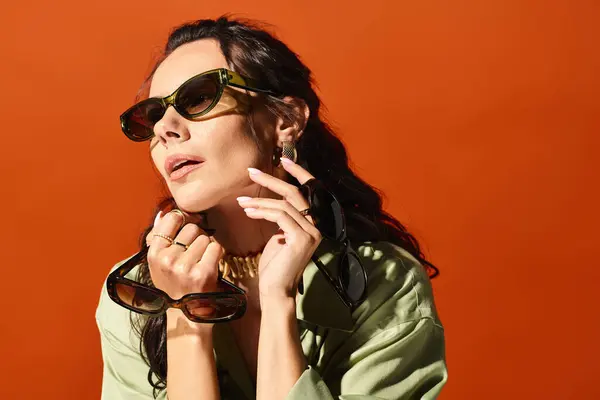 Una mujer de moda posa confiadamente en un estudio con gafas de sol y una camisa verde, exudando vibraciones veraniegas sobre un fondo naranja. - foto de stock