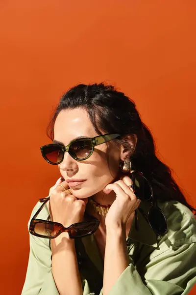 Une femme élégante portant des lunettes de soleil prend une pose confiante dans un studio sur un fond orange vif, respirant les vibrations de la mode estivale. — Photo de stock