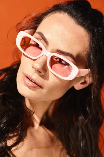 Une femme à la mode avec des lunettes de soleil rose sportif aux cheveux longs respirant les vibrations estivales dans un studio sur un fond orange. — Photo de stock