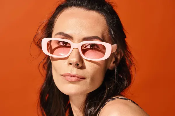 Una bella donna che indossa occhiali da sole rosa e un top nero posa in uno studio su uno sfondo arancione, in mostra la moda estiva. — Foto stock