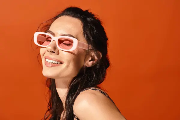 A stylish woman wearing pink sunglasses smiling in a studio against an orange background, exuding summertime fashion vibes. — Stock Photo