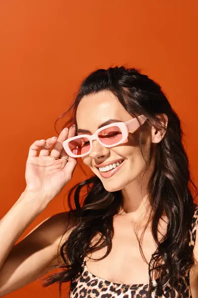 Una mujer con estilo exuda confianza en las gafas de sol de color rosa y un top estampado de leopardo sobre un fondo naranja. - foto de stock
