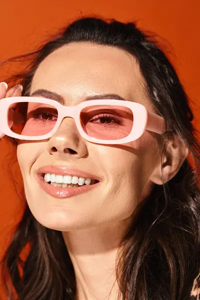 Una mujer bonita con gafas de sol rosadas sonríe brillantemente en un estudio, mostrando la moda veraniega sobre un fondo naranja. - foto de stock