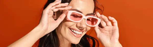 A lovely woman beams while sporting pink heart-shaped sunglasses against an orange studio backdrop. — Stock Photo